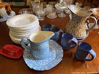 Ceramic and stoneware lot including stack of white plates, blue pitchers, blue mugs, red covered butter dish, and blue speckled serving plate.