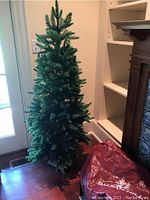 Full 5ft green artificial Christmas tree standing upright with visible metal base and nearby red storage bag with partial Balsam Hill label.