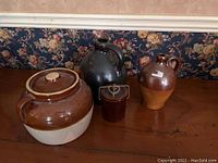 Photo showing four earthenware pottery pieces on a wooden surface against floral wallpaper. Items are a bean pot with lid, two jugs, and a small crock jar.