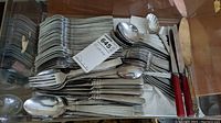 Photo showing stacked stainless steel forks, spoons, large serving spoons, and two knives with black Bakelite handles. A plastic container under the flatware is partially visible.