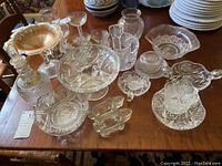 Wide view of assorted glassware pieces on wooden table including compotes, a glass vase, and decanter