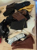 Various gloves including black leather, brown leather, yellow leather, white cotton and dark gloves stacked on a wooden table.