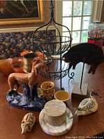 Photo showing hunting dog figurine, wrought iron fruit basket, duck figurine, stone jar with marble lid, marble wine holder, ceramic pots, and piggy bank on a table