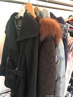 Three ladies outerwear items on hangers: black wool coat, brown alpaca wool coat with fur collar, grey ski jacket.