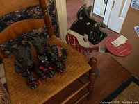 Six Scottie dog figurines placed on a woven wood seat chair plus wooden Scottie dog server resembling a stool with painted dog head and red plaid seat.