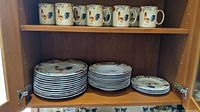 Photo of dinner plates, soup bowls, salad plates stacked on wooden cabinet shelf along with mugs on top shelf