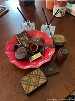 Photo showing red ceramic dish with ruffled edges holding various tartan-patterned items and two marble diffuser bottles with black reeds placed behind it on a table.
