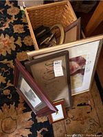 Five framed art pieces, storage basket, cast-iron match holder grouped for sale. Frames stacked and leaning inside basket and on the floor.