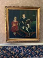 Frontal view of the oil painting showing two children, one on a rocking horse and the other standing, in a gold-toned ornate frame hung on wall above floral wallpaper.
