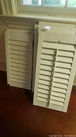 Group of white plantation shutters stacked, showing varying cuts and measurements. Shutters have horizontal louvered slats and some have visible hinges.