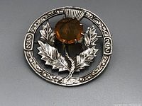 Close-up front view of antique sterling silver circular Scottish pin featuring a detailed thistle design and amber stone.