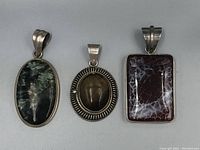 Three sterling silver gemstone pendants shown side by side: an oval green stone, an oval brown stone with silver frame, and a rectangular brown and white veined stone.