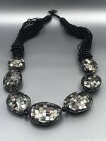 Full view of necklace showing seven large abalone mother of pearl mosaic oval beads connected by small round black beads and ending in multi-strand black cord tassels with toggle clasp.