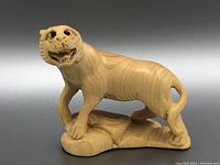 Front and side views of a wooden carved tiger netsuke showing detailed facial features, open mouth, textured base and curled tail.