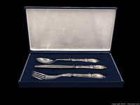 Opened navy blue textured box showing dark blue felt base holding a sterling silver spoon, knife, and fork with decorative handles inside, lid lined in white satin.