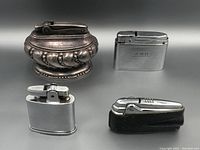 Four vintage lighters displayed against neutral background including one ornate silver-tone lighter, two polished silver-tone with/without engraving, one with leather case.