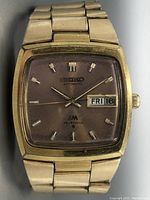 Front view showing gold-tone square watch case and faded pink dial with day/date window and Seiko branding.