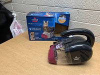 Side view of new Bissell Pet Hair Eraser handheld vacuum cleaner and its original box on a table.