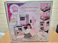 Front face of the box showing the Disney Princess Style Gourmet Smart Kitchen Playset with images of a child playing and features like stove recognizing pots and pans, talking phrases, and included accessories.