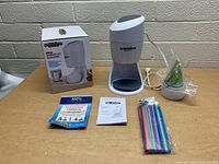 White Hawaiian shaved ice machine on a tabletop with original box, instruction manual, colored straws in plastic wrapping, and small cups with lids in sealed packaging.