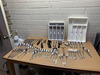 Photo of large assortment of flatware and kitchen utensils on table showing forks, spoons, knives, corkscrews, vegetable peeler, bottle opener, and plastic utensil trays.