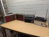 Five vintage portable radios arranged side by side on a wooden table in a room with light-colored brick walls.