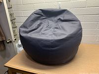 Dark gray fabric bean bag chair shown from an angle on a wooden surface against a white brick wall.