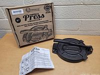 Victoria brand cast iron tortilla press with original box and instruction manual on a wooden surface against a beige brick wall.