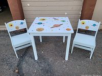 Front view of the white wooden kids table with space-themed top and two matching chairs showing the space design on backrests.