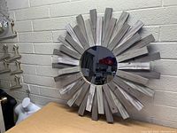 Front and angle views of the round wall mirror with a rustic wooden sunburst frame on a beige brick wall and tabletop.