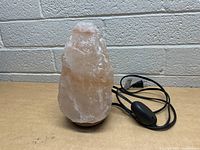 Himalayan salt lamp showing rough pink salt crystal with attached black electrical cord and inline switch, mounted on wooden base.