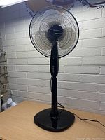 Full view of black oscillating pedestal fan placed on a table against a white brick wall.