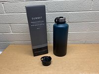 Photo of boxed Simple Modern Summit 84 oz insulated water bottle in dark teal to black gradient, bottle with screw-on lid and extra lid on table against brick wall.