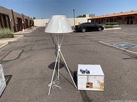Threshold white tripod floor lamp standing outdoors next to the packaged box containing the lamp accessories and bulbs.