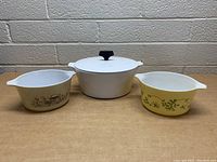 Three vintage kitchenware pieces against brick wall background on wood surface: white Corning Ware pot with lid in center, beige Pyrex bowl with countryside design left, yellow Pyrex bowl with floral design right.