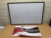 VIZ-PRO whiteboard 35x24 inches with black aluminum frame resting vertically against a brick wall on a wooden surface, original packaging placed at front.