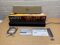 Sound bar, remote wrapped in plastic, power cable in plastic, user manuals, and boxes displayed on table.