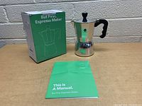 Photo showing the espresso maker, original green box labeled 'But First, Espresso Maker,' and the green user manual on a wooden surface against a white brick wall.