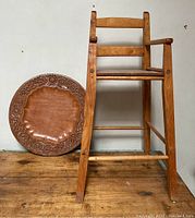Full view of antique child's wooden chair and round carved wood platter side by side on wooden surface.