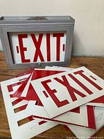 Photo of metal exit sign housing and multiple red 'EXIT' faceplates stacked in front of it on a wooden surface.