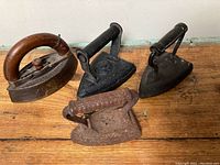 Top view showing four antique irons on wooden surface, various handle types and rust patina visible