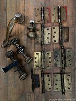 View of entire lot showing multiple brass door hinges in different sizes and finishes, two sets of ribbed brass door knobs, a curved brass door handle, and a round brass doorknob.