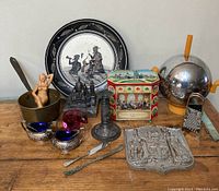 Photo showing entire collection of vintage home decor and kitchen items arranged on wooden surface. Items include decorative plate, ceramic figure, metal sculptures, glass art pieces, tin box, and small kitchen tools.