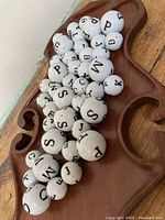 Photo showing multiple white ceramic balls with black alphabet letters arranged on a wooden tray. Various sizes and letters visible.