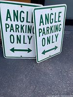 Two heavy metal angle parking signs with green lettering and directional arrows on white background. Both signs read 'ANGLE PARKING ONLY'.