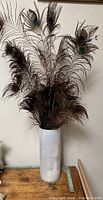Front view of tall white glass vase with natural peacock feathers inside, on wooden surface against neutral background.