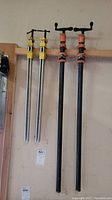 Photo showing four wood clamps: two yellow quick-release clamps with silver bars and two older black pipe clamps with orange handles, hung on a wall.
