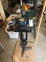 Photo shows the full bench grinder unit mounted on pedestal stand with attached lamp, surrounded by garage storage items.