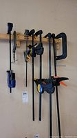 Photo showing 8 clamps on a wall, including 2 metal C clamps and 6 bar clamps, viewed from distance