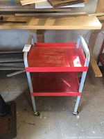 Side view of the two-tiered metal push cart showing red shelves and gray metal frame with wheels, dusty and in used condition